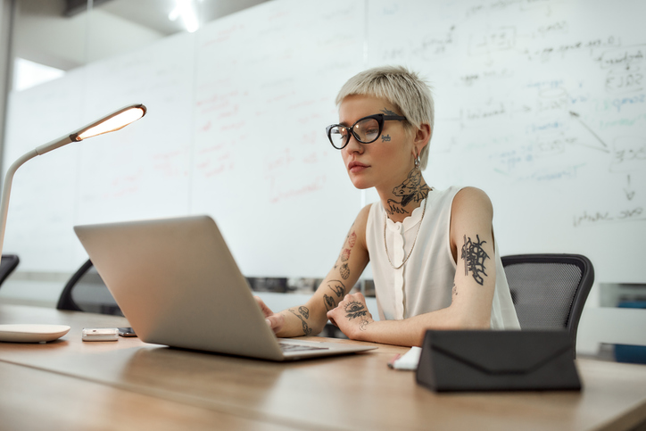 Young tattooed woman with short haircut using laptop while sitting at her working place in the meeting room